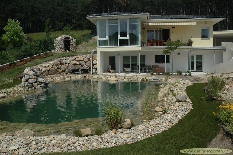 Swimming pond with boulder edges and a planted shelf below a hillside home, Austria